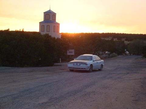 1990 Ford Taurus - A white sedan is parked on a dirt road with a tower in the background during sunset.