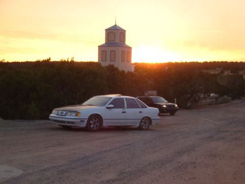 1990 Ford Taurus - A white 1990 Ford Taurus sedan and a dark SUV are parked near a building at sunset.
