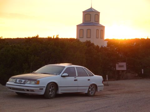 1990 Ford Taurus - A white 1990 Ford Taurus with a black hood sits parked in front of a tower during a sunset.