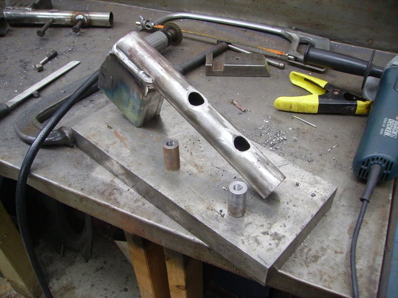 Fabricated steel tube arm with drilled holes and two bushes on a workbench with grinder and clamps