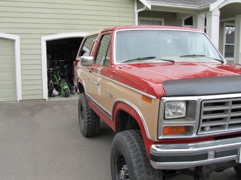 1984 Ford Bronco - Red and tan Ford Bronco with lifted suspension parked in front of a garage.