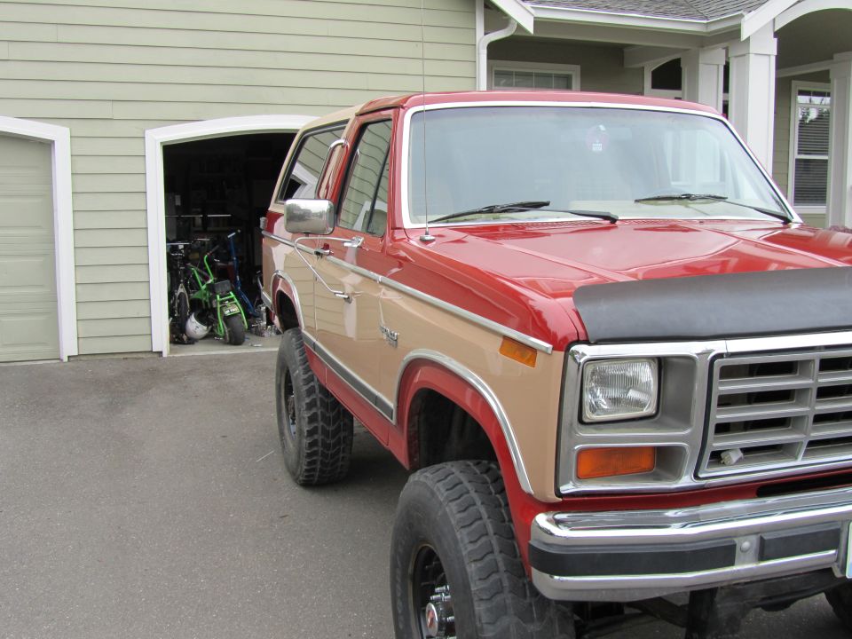 Red and tan 1980s Ford Bronco parked in driveway, front quarter view. Garage in background