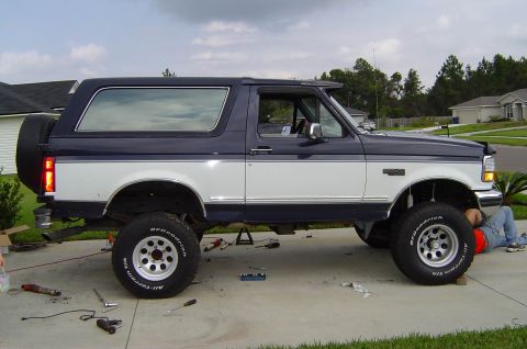 1993 Ford Bronco - A person works on the front wheel of a lifted two-tone Ford Bronco on a suburban driveway.