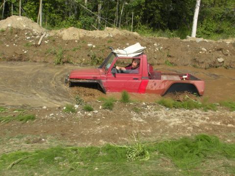 1987 Suzuki Samurai - A red off-road vehicle navigates a muddy terrain, with its driver visible behind the wheel.