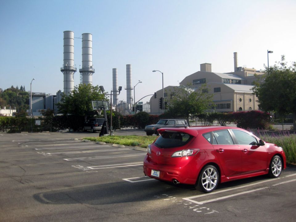 Red hatchback parked in a lot with a power plant in the background. Another car is parked nearby