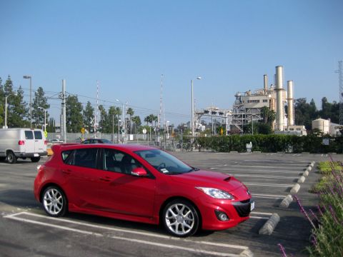 1989 Mazda MX-6 - A red hatchback is parked in a lot with a white van driving by and an industrial complex in the background.