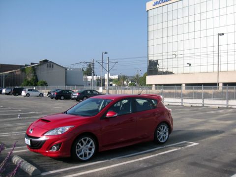 1989 Mazda MX-6 - This red hatchback, likely a Mazda3, is parked in a lot with other vehicles and a large building in the background.