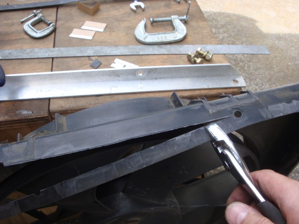 Black plastic radiator fan shroud with a metal tool being used on its edge, resting on a wooden surface with clamps and bolts