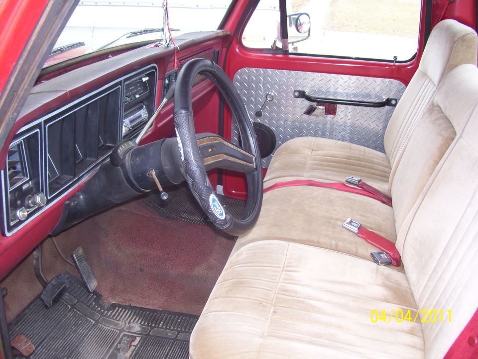 1978 Ford F-100 interior showing dashboard, steering wheel, and tan seats with red seatbelts. Diamond plate door panel