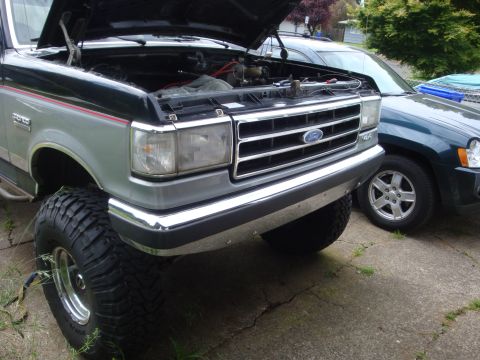 1989 Ford Bronco - A lifted Ford Bronco with its hood open is parked next to a dark green SUV.