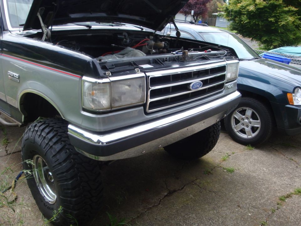 Two-tone silver and black Ford Bronco with hood open, chrome bumper, and large tires parked next to a dark green SUV