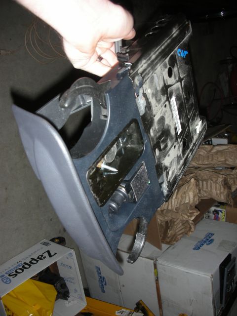 1998 Ford Crown Victoria - gray plastic center console with an open cupholder is held up, its black underside marked CWT, above cardboard boxes in a storage area.