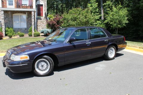 1999 Ford Crown Victoria - A dark blue sedan is parked on an asphalt surface in front of a residential building.