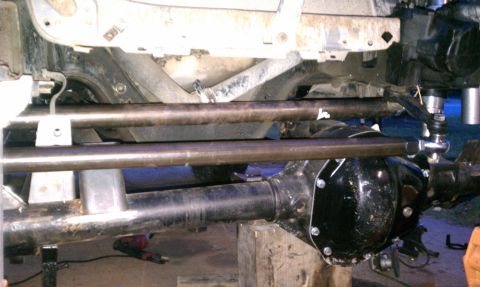 1990 Ford Bronco - Underbody view of a solid front axle with a black cast differential housing resting on a wood block, with two long bare-steel radius/link t…