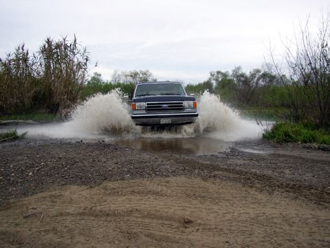 1989 Ford Bronco - A dark blue Ford F-150 truck drives through a water-filled road, creating large splashes.
