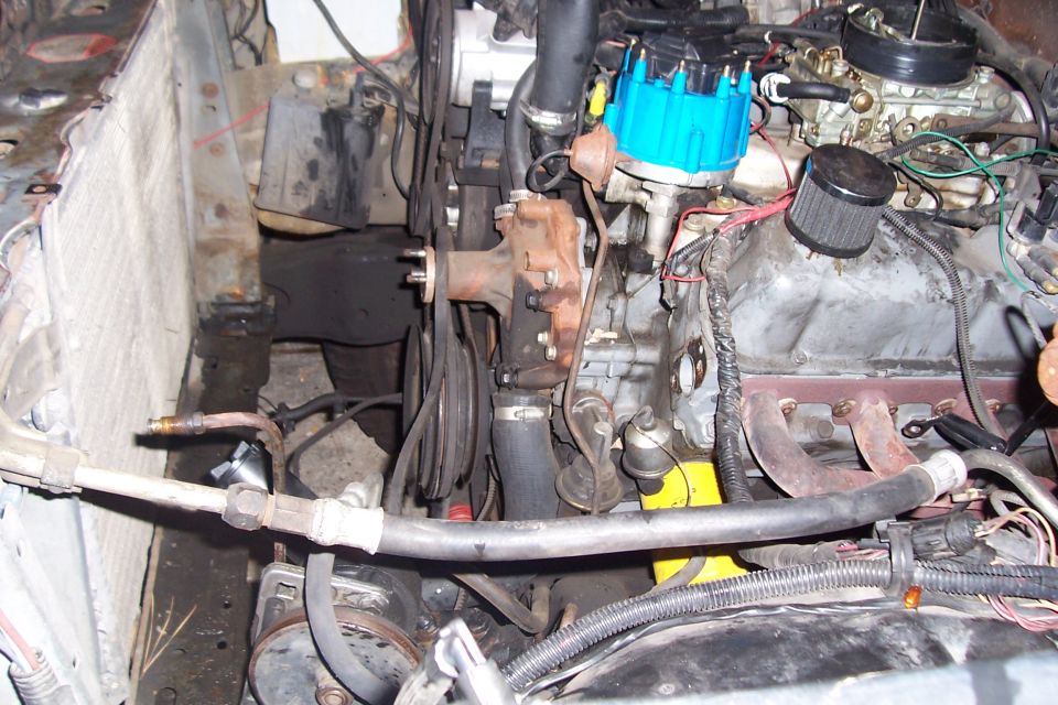 Engine bay with a blue distributor cap, carburetor, and exhaust manifold. Some hoses and wires are visible