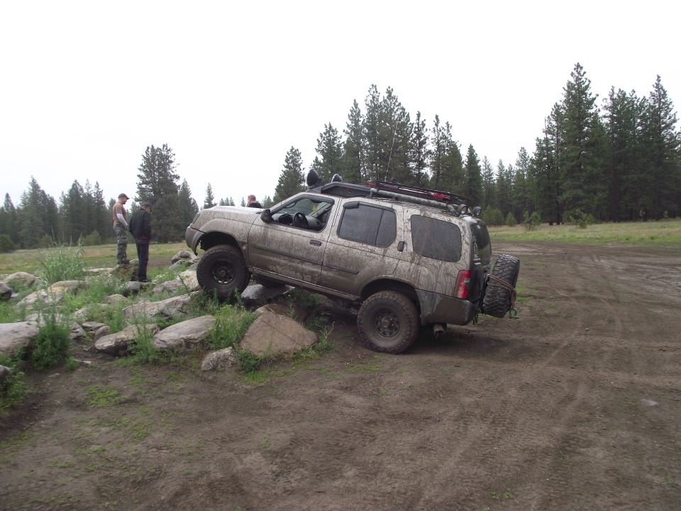 Muddy Nissan Xterra SUV climbing rocks off-road in a forest clearing, side view, front end raised