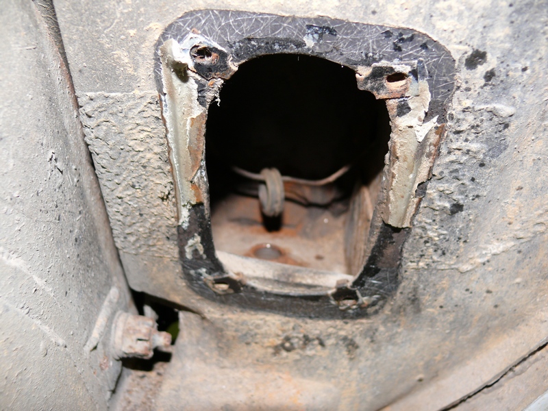 Underside of a vehicle showing a rusted, open access panel with a wire loop inside