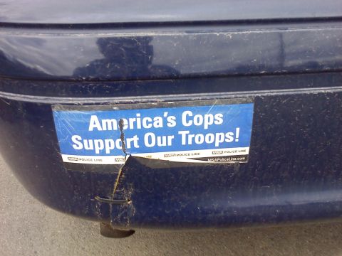 2002 Ford Crown Victoria - A close-up of a damaged bumper sticker on a dark blue vehicle reads "America's Cops Support Our Troops!"