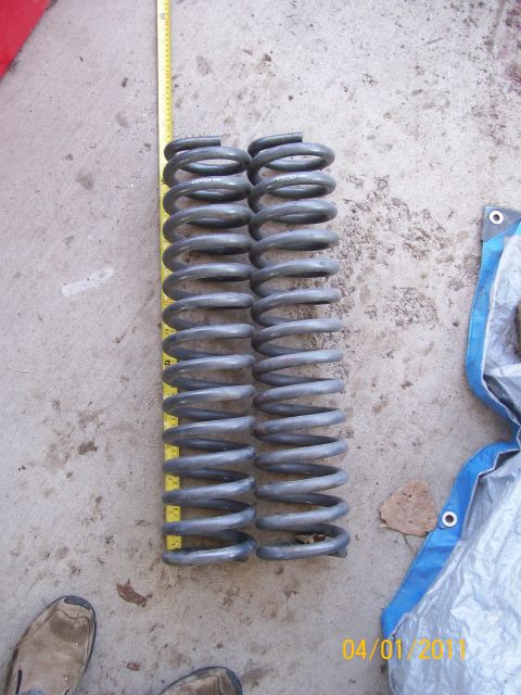1970 Ford Bronco - Two gray coil springs are laid out next to a yellow measuring tape on a concrete surface.