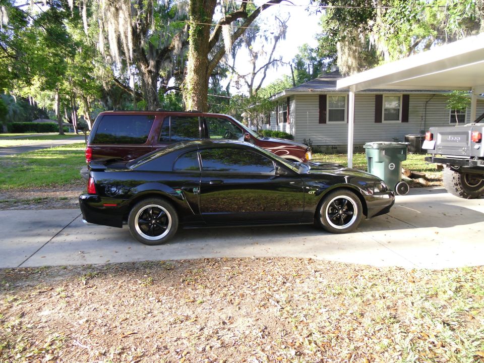 Black Ford Mustang coupe parked on driveway, side view. Another SUV and a Jeep are also visible
