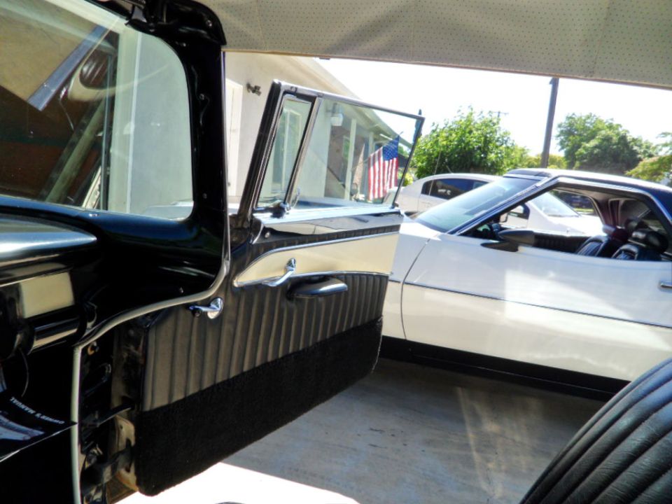 Black and tan interior of a 1956 Ford Fairlane with the driver's door open, showing a white car parked alongside