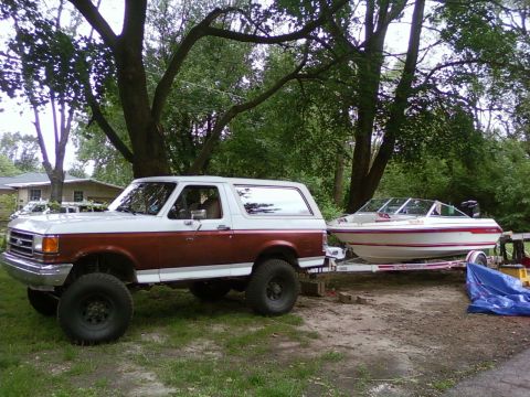 1989 Ford Bronco - A two-tone 1989 Ford Bronco is parked on a dirt lot, hitched to a boat on a trailer.