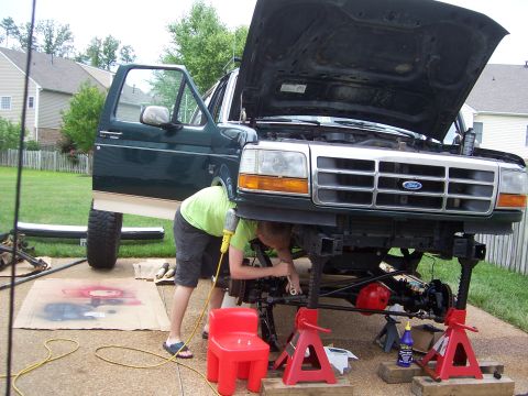 1992 Ford Bronco - A person works on the front suspension of a green 1992 Ford Bronco lifted on jack stands in a driveway.