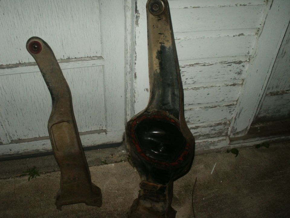 Two rusty automotive suspension parts, one with a dark circular opening, lie on concrete in front of a white paneled door