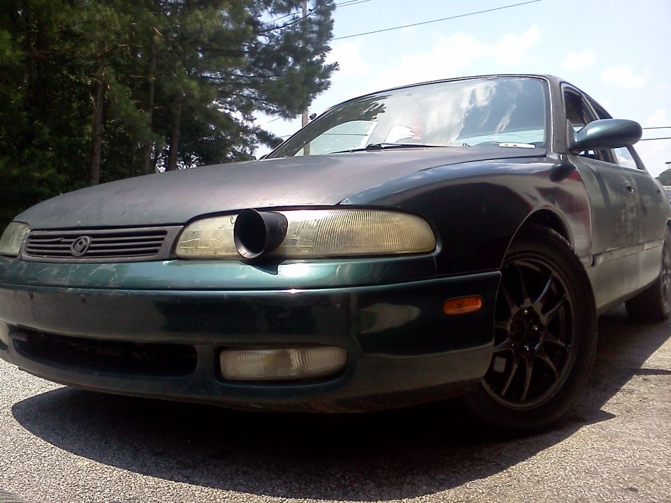 Faded green Mazda sedan with intake tube through headlight and black alloy wheels