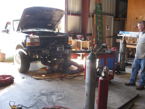 1994 Ford Bronco - Mechanic welding on the underside of a Ford Bronco in a garage with tools and equipment around.
