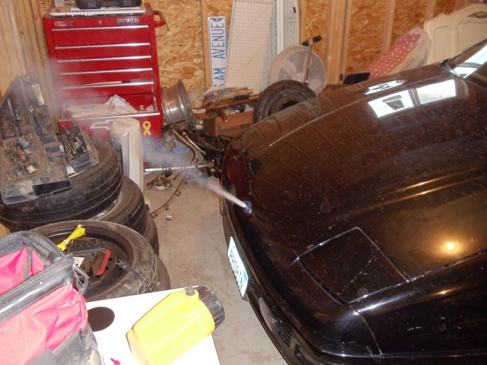 Black vehicle hood being spray-painted in a wood-framed garage with stacked tires