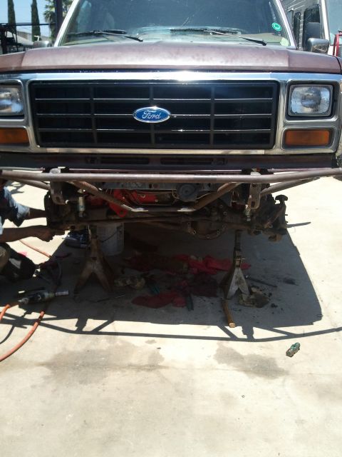 1982 Ford Bronco - The front end of a brown Ford Bronco is disassembled and supported by jack stands, revealing the engine and suspension components.