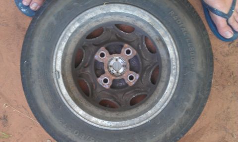 1991 Ford Festiva - Overhead shot of an old four-lug slotted steel wheel lying face-up on red dirt, the center and lug area heavily surface-rusted with a bare…