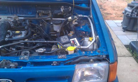 1991 Ford Festiva - The engine bay of a blue vehicle is shown, the battery and various hoses. A Supermotors.net logo is present.