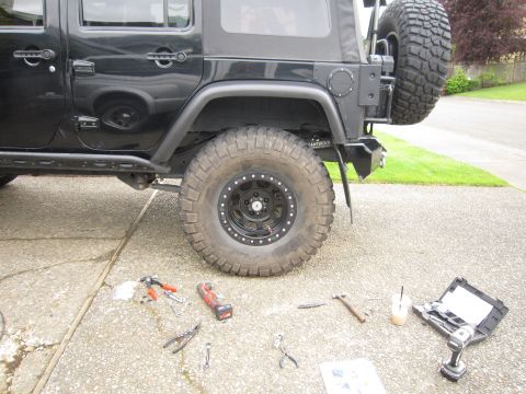 2008 Jeep Wrangler - Driver's-side rear quarter of a lifted black four-door Jeep Wrangler in a driveway mid-project, with a mud-terrain tire on a black beadlock…