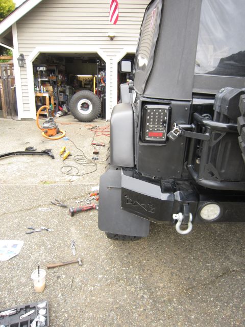 2008 Jeep Wrangler - Rear corner of a black Jeep Wrangler in a driveway during a bumper install, showing a steel rear bumper with a D-ring shackle and round LED…