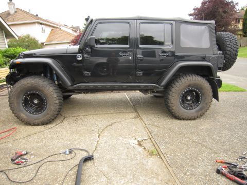 2008 Jeep Wrangler - Full driver's-side profile of a lifted black four-door Jeep Wrangler with a black soft top, parked on a suburban driveway. It rides on mud-…