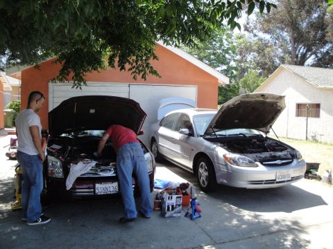 2003 Ford Taurus - Two men work on cars in a driveway, one a silver 2003 Ford Taurus with its hood open.