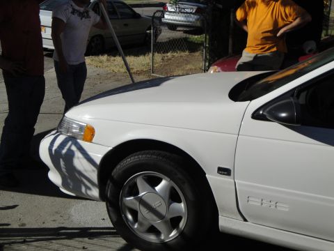 2003 Ford Taurus - A white 2003 Ford Taurus sedan is parked outside with people standing around it.