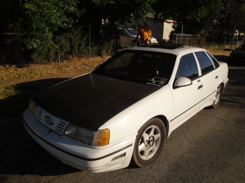 2003 Ford Taurus - A white sedan with a black hood and a sunroof is parked on the street. A person stands near another vehicle in the background.