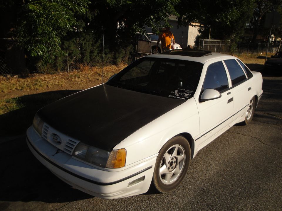 White sedan with a black hood and sunroof parked on a street. Another car and a person are visible in the background