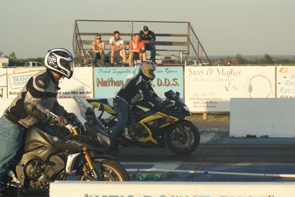 Two motorcycles and riders at the starting line of a drag strip, with spectators in the background