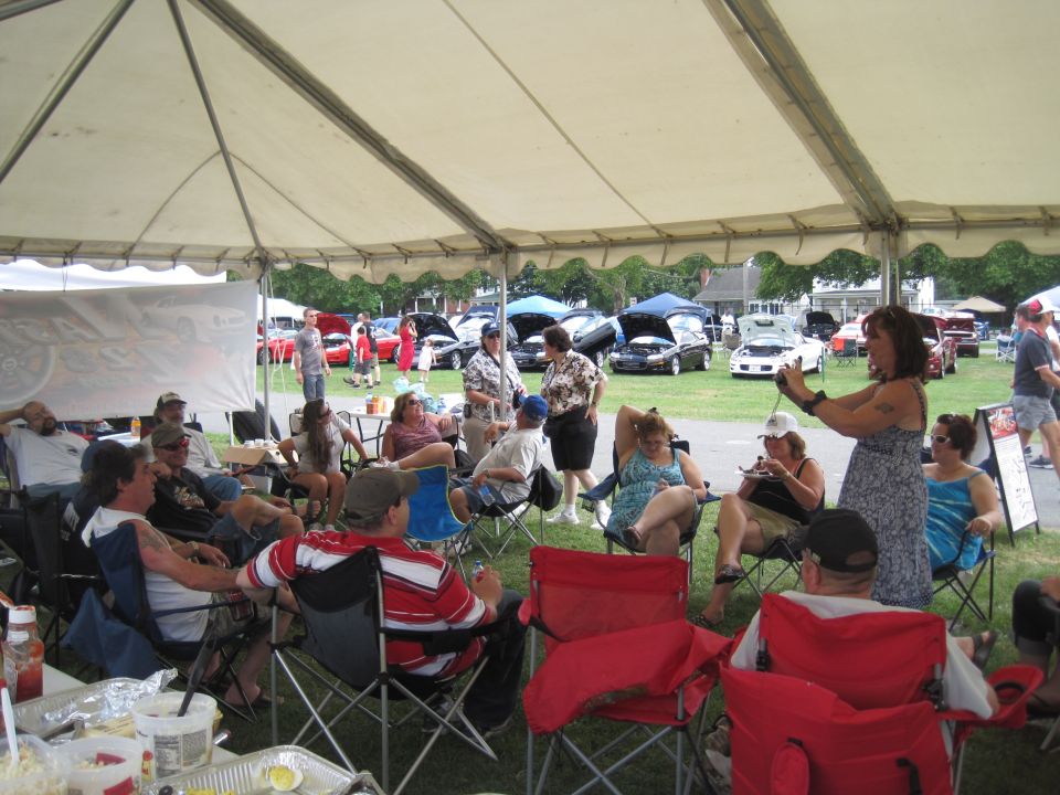 People sitting under a tent at a car show, with cars displayed in the background