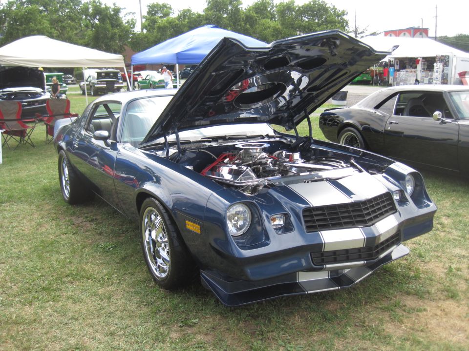 Blue muscle car with silver racing stripes, hood open showing engine. Another black car is parked nearby