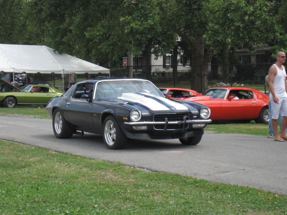 Dark blue 1970s Chevrolet Camaro with white racing stripes on hood and roof, driving on asphalt. Red Camaro and green Camaro visible in background