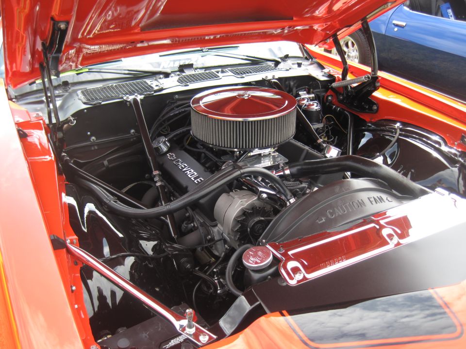 Orange car's engine bay with a Chevrolet engine, chrome air cleaner, and fan shroud. Blue car visible in background