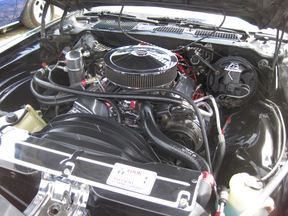 Engine bay with chrome air cleaner and V8 engine components. A blue car is partially visible on the left