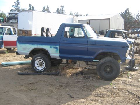 1978 Ford Bronco - A blue 1978 Ford Bronco sits in a junkyard, showing primer on its rear quarter panel and two seats in the open bed.