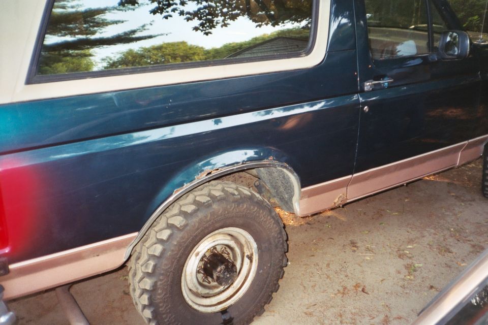 Rear quarter of a teal Ford Bronco with cut fender lip and mud tire on a white steel wheel